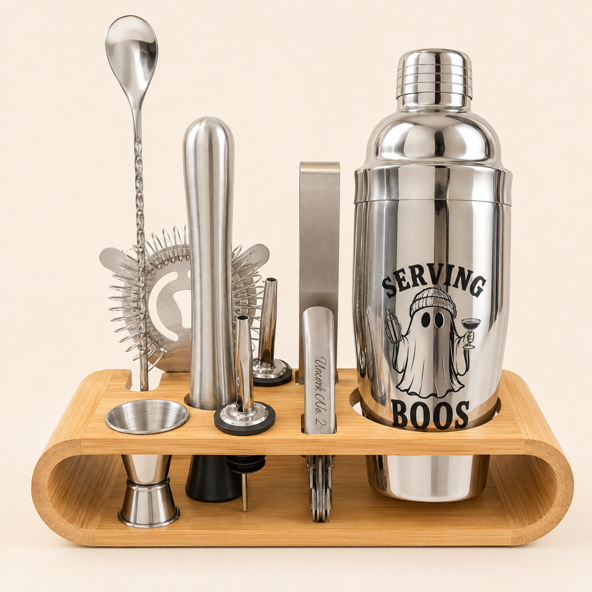 A stainless steel cocktail bar tool set arranged on a curved bamboo holder against a light beige background. The set includes a shaker tin with a ghost graphic and the text 'SERVING BOOS', a muddler, strainer, jigger, spoon, and other bar accessories.